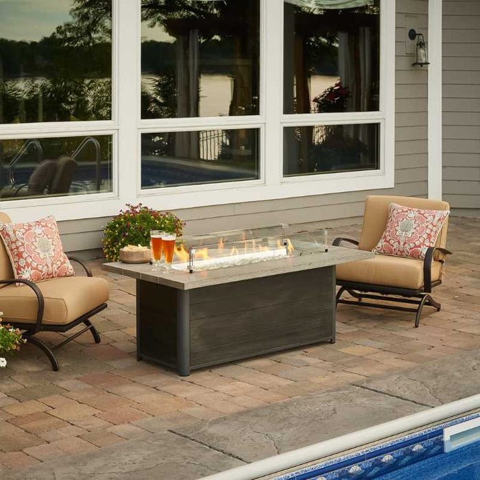 The Outdoor Greatroom Cedar Ridge Linear Gas Fire Pit Table near Pool with Clear Tempered Fire Glass Gems, Fire Burner On and Glass Wind Guard