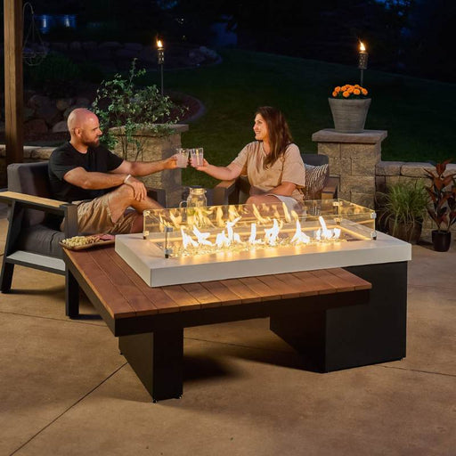 The Outdoor Greatroom Iroko Uptown Linear Gas Fire Pit Table Installed at Terrace with Happy Couple and Clear Tempered Fire Glass Gems plus Fire Burner On and Glass Wind Guard Installed