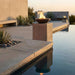 Warming Trends AON Aura concrete charcoal fire bowl with natural gas flames on a concrete pedestal by a modern luxury swimming pool at sunset.