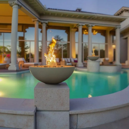 Warming Trends AON Aura concrete fire bowl in color sand with active flames, placed on a stone pillar alongside a luxury swimming pool area.