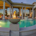 Warming Trends AON Aura concrete fire bowl in color sand with active flames, placed on a stone pillar alongside a luxury swimming pool area.