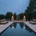 Warming Trends AON Ember square concrete fire bowls in color white with active flames, elevated on matching stone pillars both sides of a luxury rectangular swimming pool area at dusk.